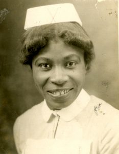 Black and white image of young black nurse looking into the camera. She wears a white nurse's hat and a white collared nurse's uniform.